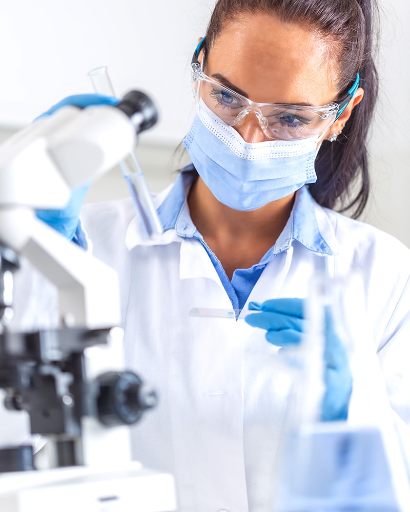 Woman in white coat, protective goggles and face mask looks at test-tube